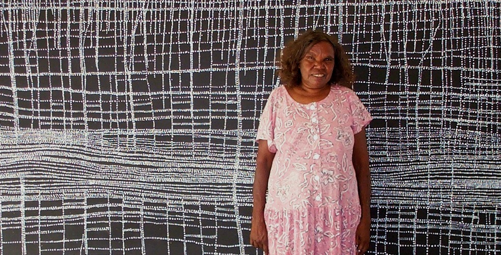 The artist Dorothy Napangardi standing in front of a large black and white painting of Mina Mina ceremonial grounds.